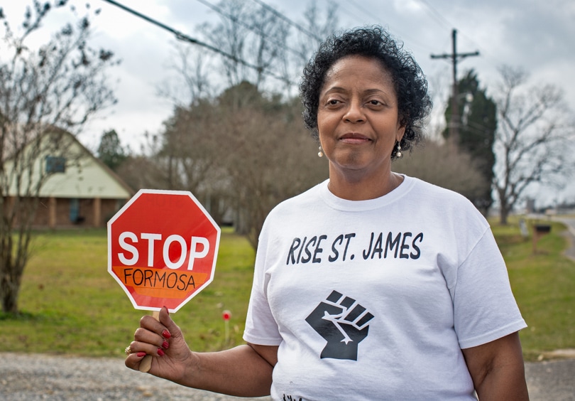 Sharon Lavigne stands outside holding a small stop sign and wearing a white t-shirt reading 'RISE ST. JAMES' and a closed fist