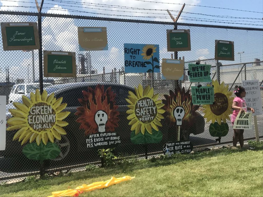 Hand painted posters and murals of sunflowers and skulls remembering community members lost to pollution line a chainlink fence outside the PES Refinery.