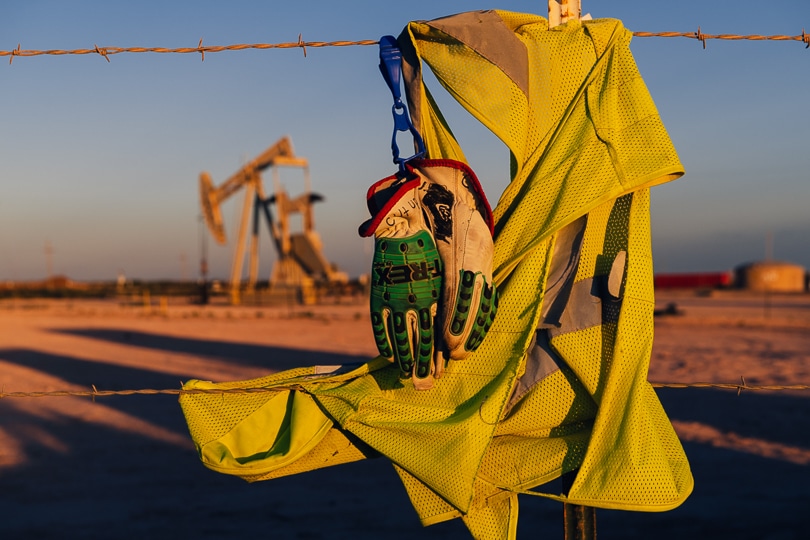 A laid-off oilfield worker's vest and gloves hang on a fence post in front of an idled pump jack in Eddy County, New Mexico at sunset.