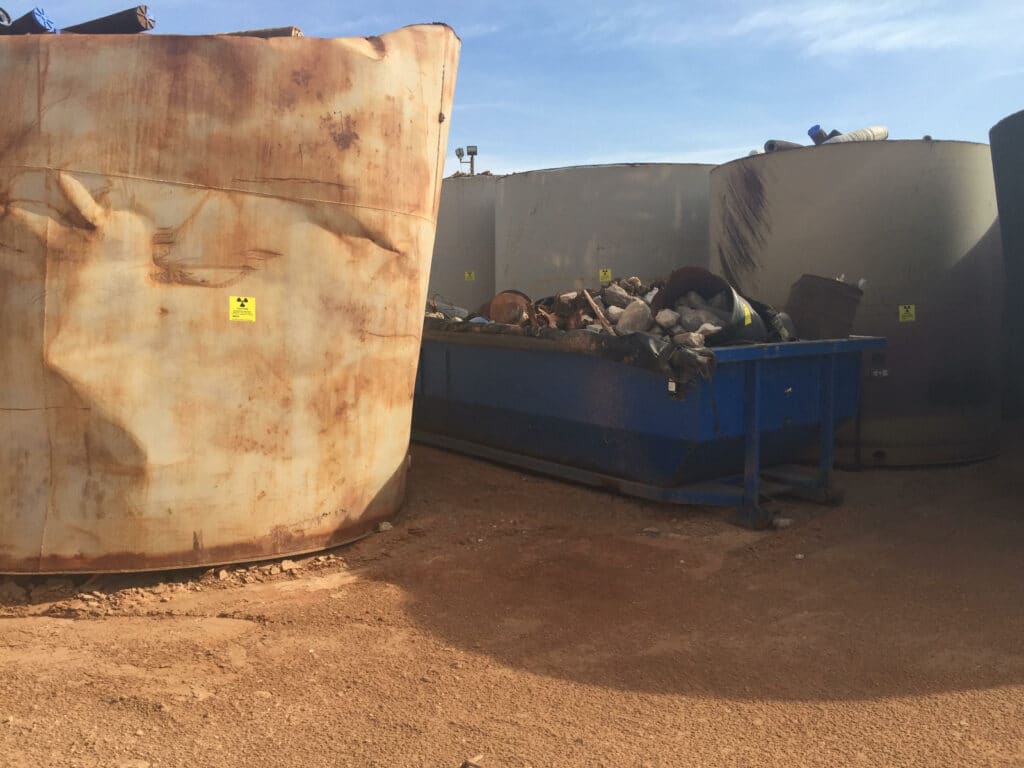 Large rusted and dented white tank next to a blue dumpster with piles of debris and white tanks with pipe sections visible, all sitting on red-dirt desert ground