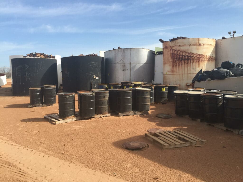 Black barrels on wooden pallets by large rusted white and blue tanks with pipe sections sticking out the top, on a red-dirt desert ground