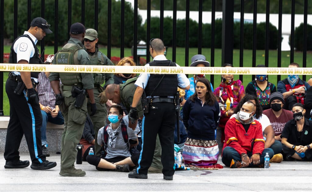 Group of seated climate protesters confronted by police by White House fence behind yellow do-not-cross tape