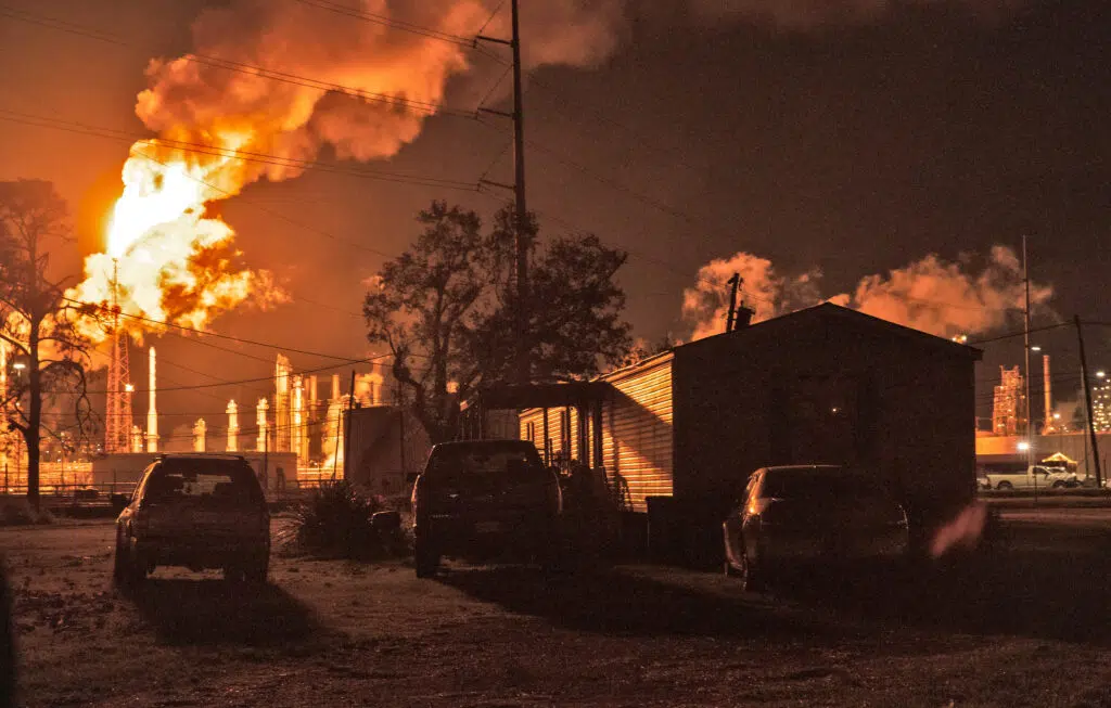 Night scene of three cars in shadow by a trailer home and orange smoke and smokestacks lit up at an oil refinery