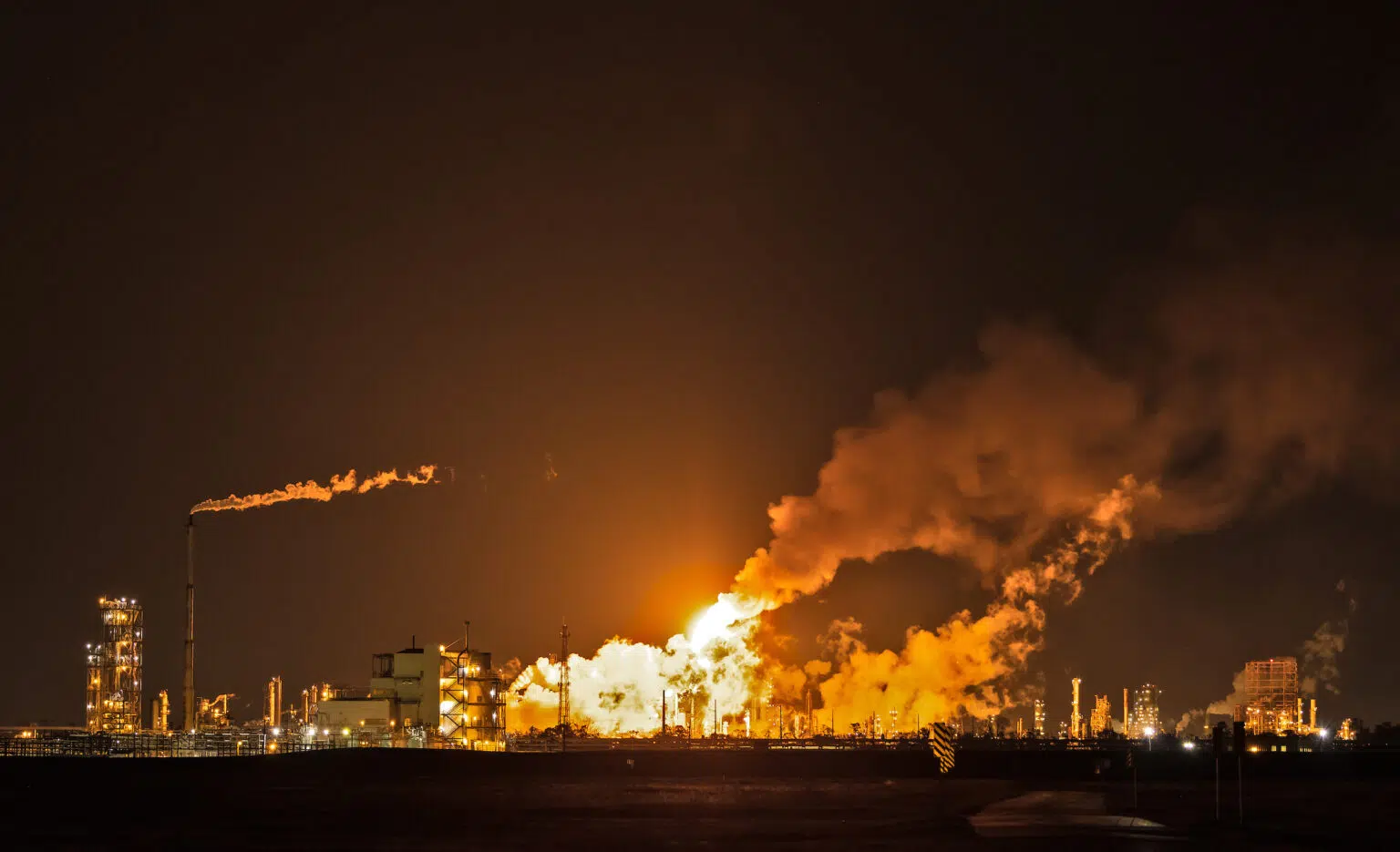 Night scene of an oil refinery with orange flares and smoke pouring out of smokestacks