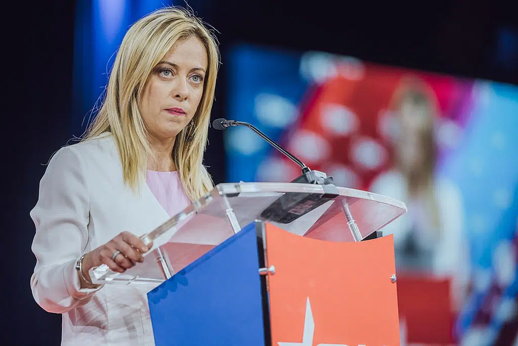 Giorgia Meloni, a blond woman in pink and white suit, looks over a clear plastic, blue, and orange podium, with her image on a jumbotron behind her.