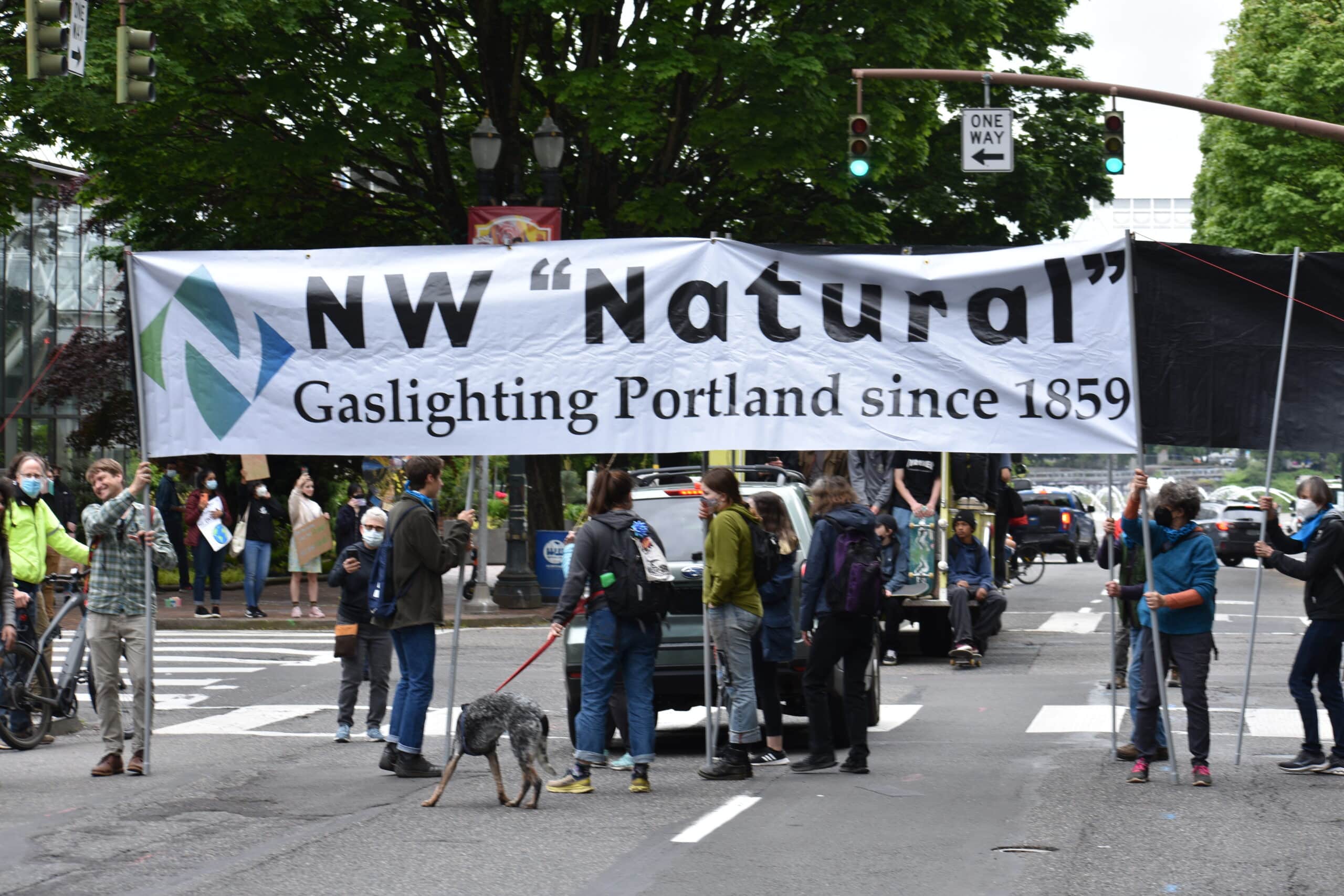 Protesters in a crosswalk hold a white banner with black writing that says "NW 'Natural' Gaslighting Portland since 1859"