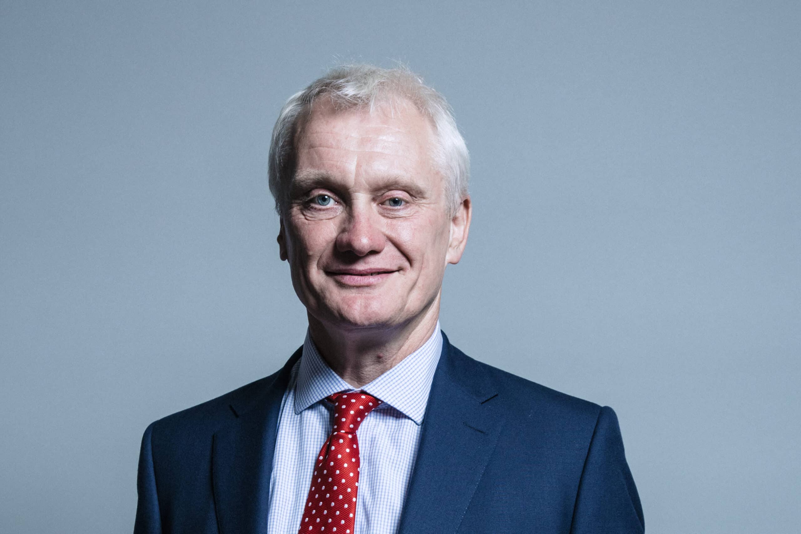 a photo of an older white man in a navy suit, blue shirt, and red tie with white polka dots