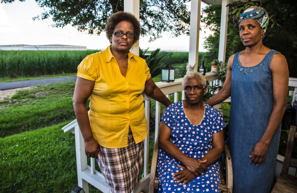 Eve Miller stands in a yellow button down shirt next to her seated mother and standing sister on her front porch in St. James, Louisiana, across from oil storage tanks in a green field.