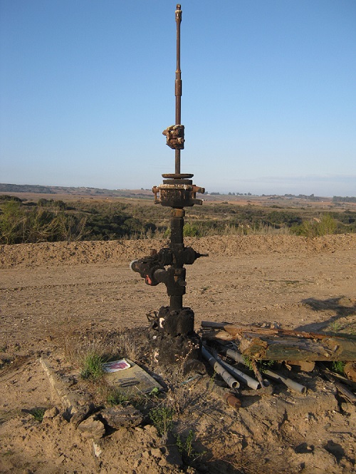 A tall rusty brown metal pipe with valves juts out of the dry soil, with a road, arid landscape, and blue sky beyond.
