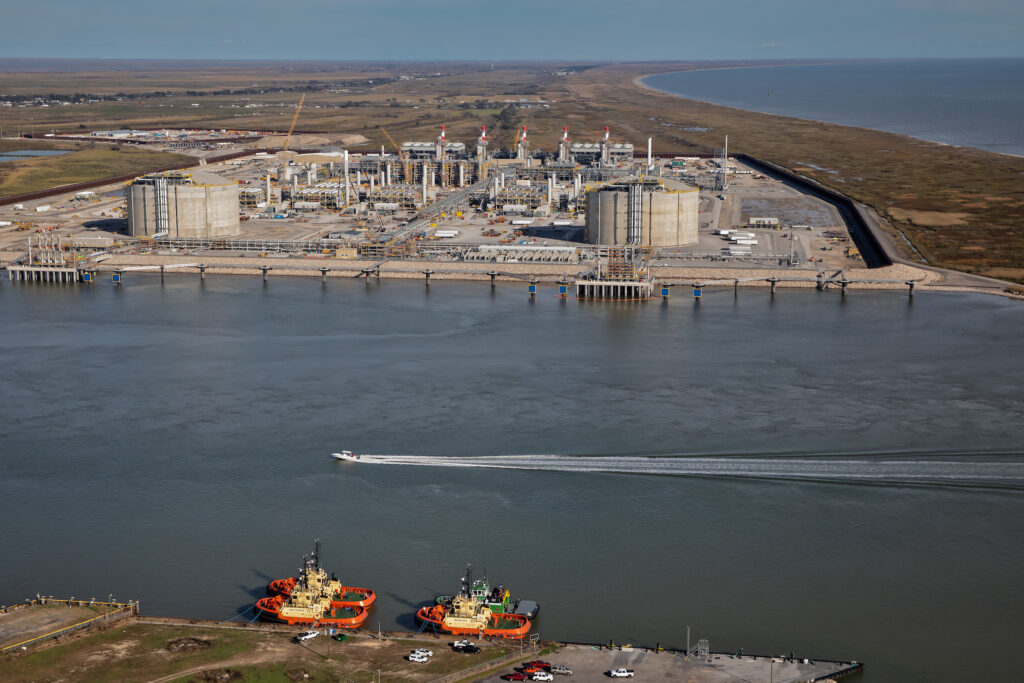 The infrastructure of the LNG terminal, with storage facilities and docks, is shown alongside the water. A small boat is passing by.