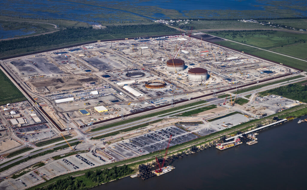 The gray and white infrastructure of the LNG complex, viewed from above, is surrounded by green open space and deep blue water.