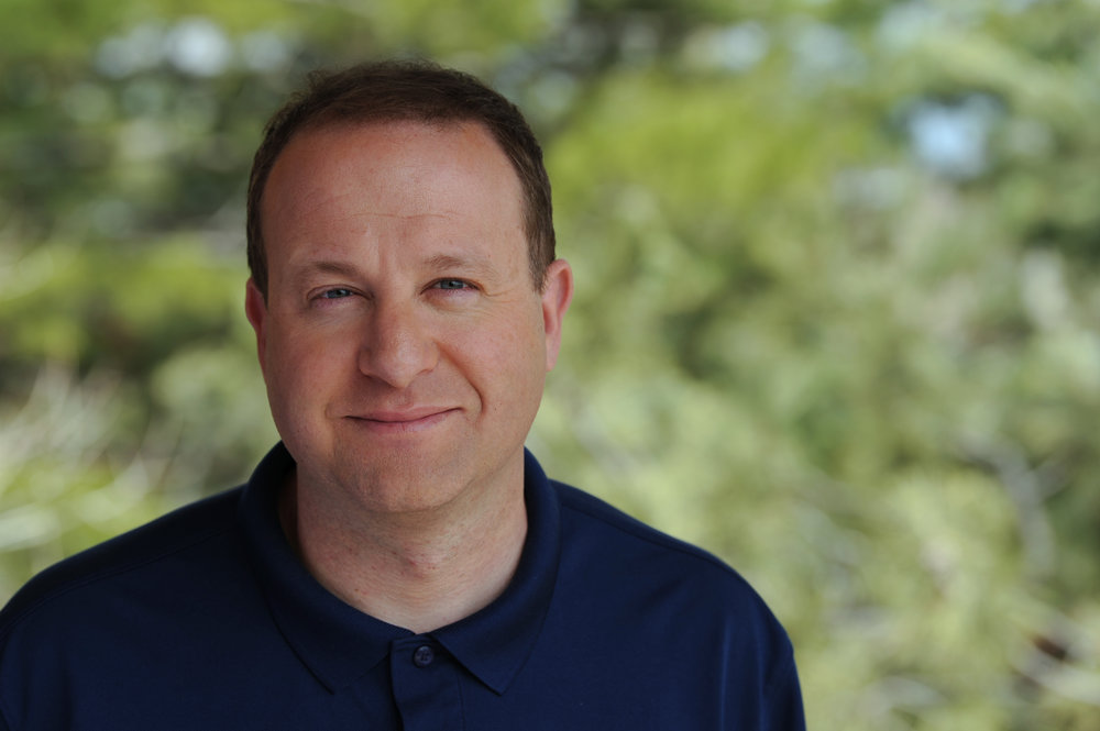 Jared Polis is smiling at the camera, blurred trees behind him. He's wearing a dark blue polo shirt.