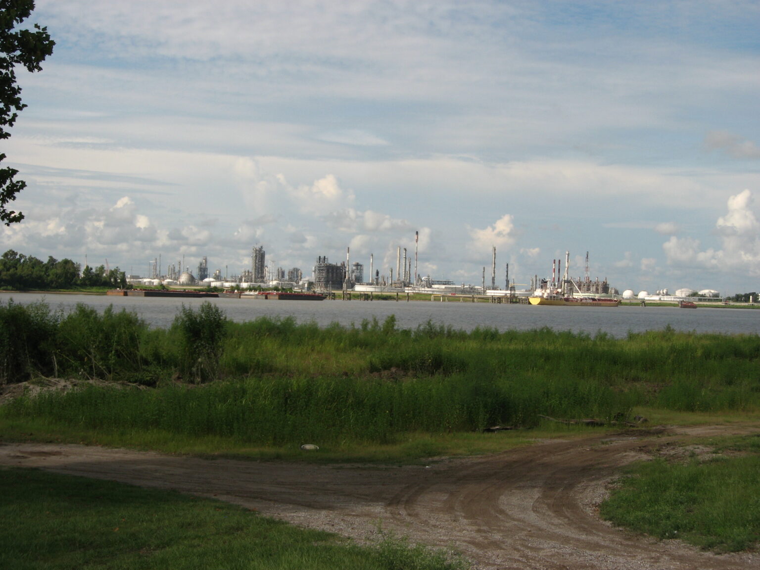 Petrochemical stacks and other elements of the industry, including ships, are viewed across the river from a spot with a dirt road.