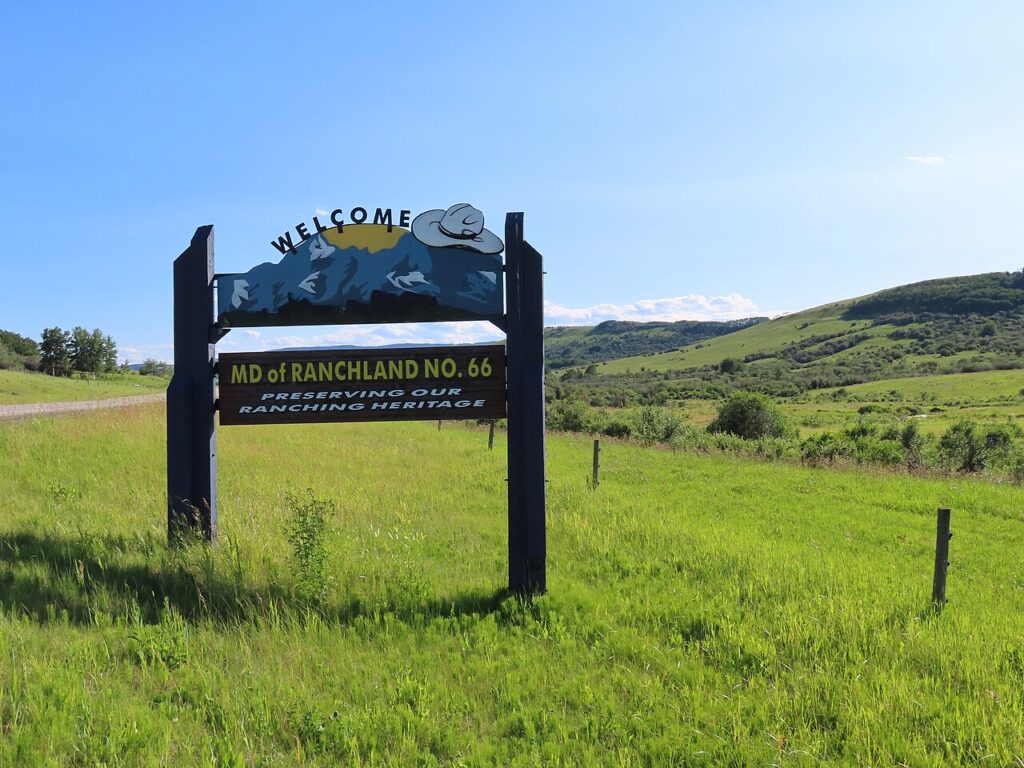 In the background are green rolling hills. In the foreground, a sign that says "Welcome" beside a cowboy hat, and below that, "MD of Ranchland No. 66, preserving our ranching heritage"