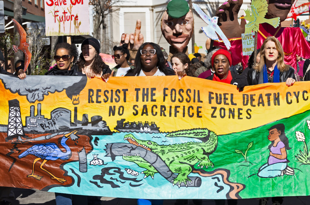 A diverse group of protesters march in New Orleans with signs and a painted banner that reads 'resist the fossil fuel death cycle, no sacrifice zones' and shows images of fossil fuel industry smokestacks polluting air and a water and an alligator tearing a pipeline in its mouth.