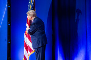 Donald Trump peers around a flag next to a blue stage curtain