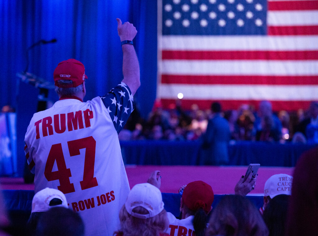 A Trump supporter wears a red ball cap and shirt that reads "Trump 47" in a crowd with a giant American flag in the background.