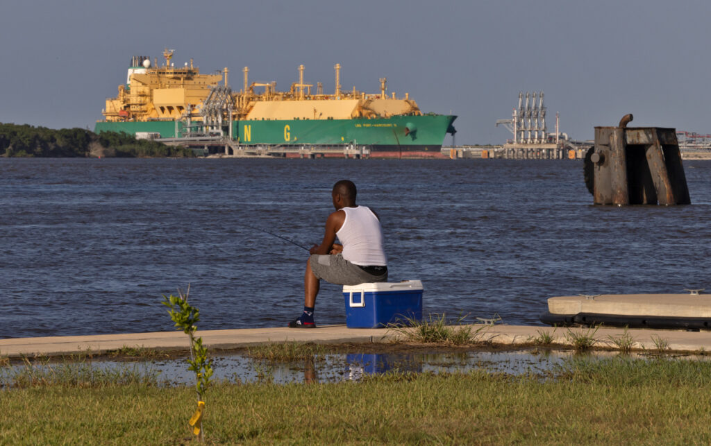 Man sitting on a cooler fishing with a large LNG tanker in the background.