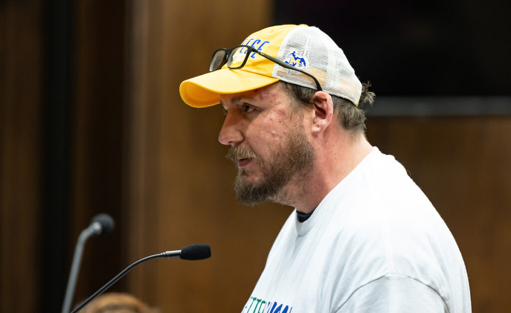 James Hiatt, wearing a white t-shirt and yellow McNeese ball cap with glasses on his head, looking left while speaking at a microphone.