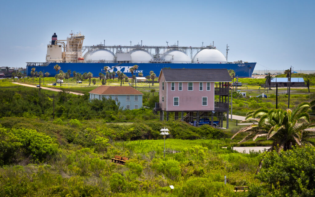 A massive tanker leaving Freeport LNG terminal, with brightly colored houses and lush vegetation in the foreground.