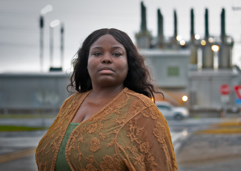 Roishetta Sibley Ozane wearing a rust-colored lace jacket over a sea-green tank top, stands in front of a chemical plant's smokestacks in the distance.